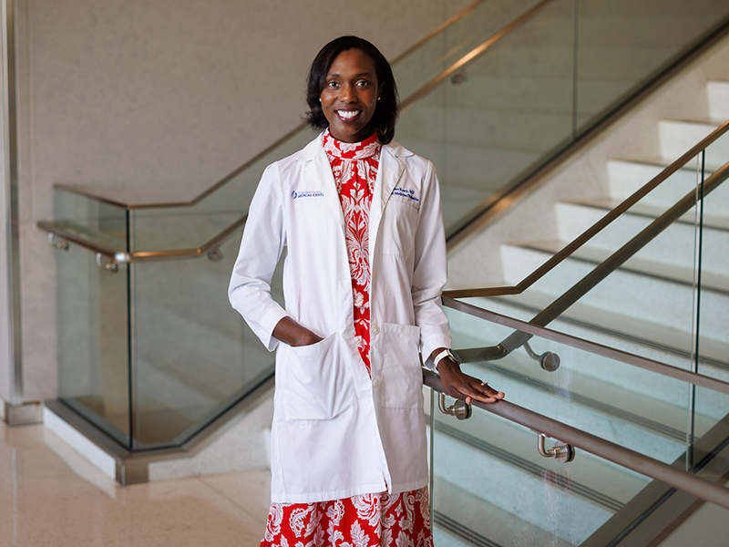 Wearing a white coat, Dr. Jasmine Kency poses in front of a starcase in the UMMC School of Medicine.